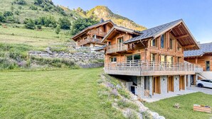 Een houten chalet met balkon, gelegen in een grasrijke omgeving met een berg op de achtergrond.