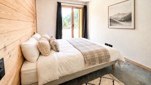 Een slaapkamer met een houten hoofdbord, een bed met witte lakens en een geruite plaid, een lijst met een landschap en uitzicht door een raam met gordijnen.