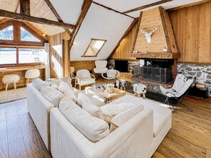 Ein gemütliches Loftsesselzimmer mit einer großen Couch, Holzbalken, einem Steinkamin und einem aufgehängten Hirschkopf.