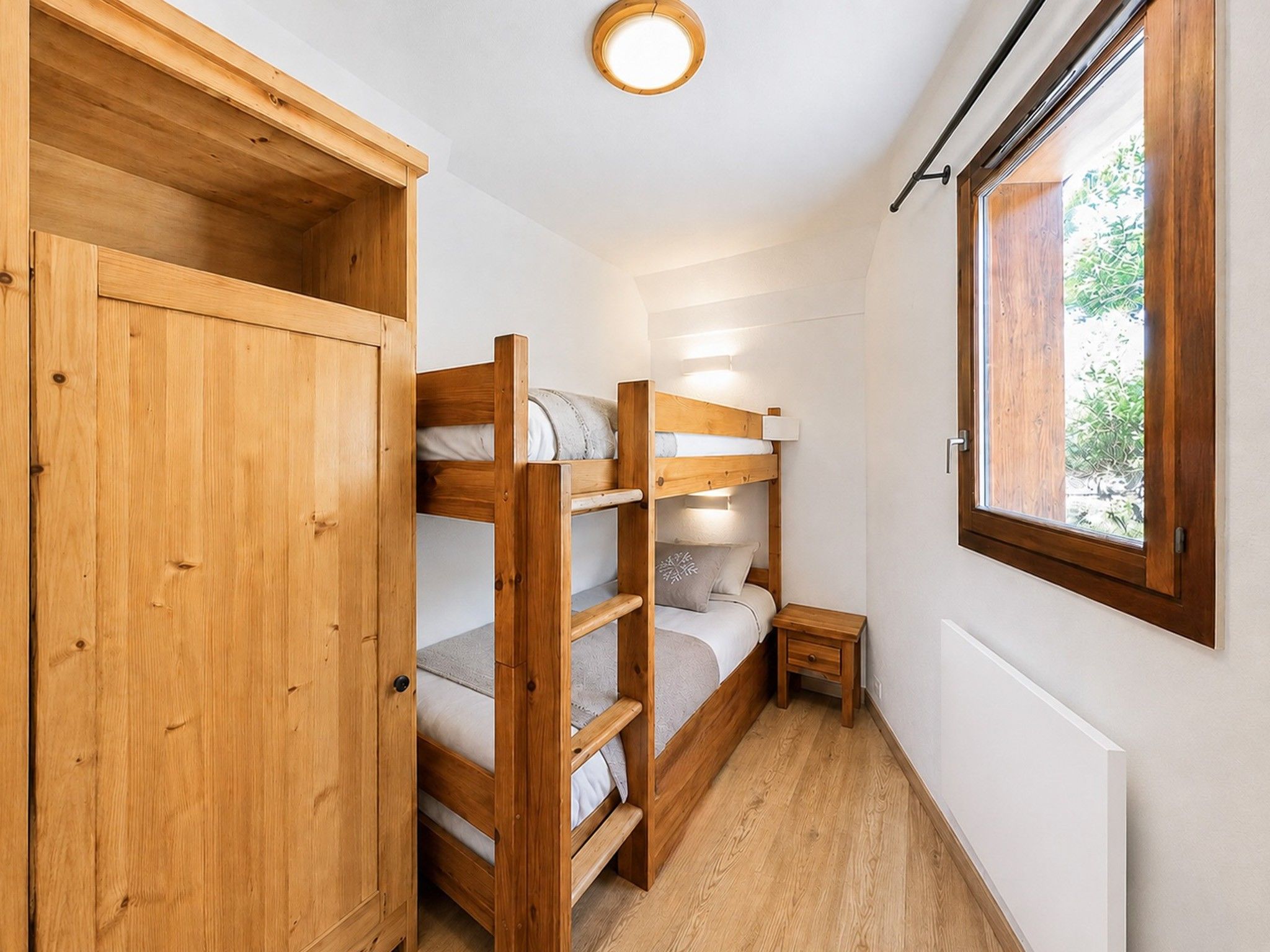 Habitación compacta con literas, un armario de madera, una mesita de noche pequeña y una ventana con vistas al verdor.