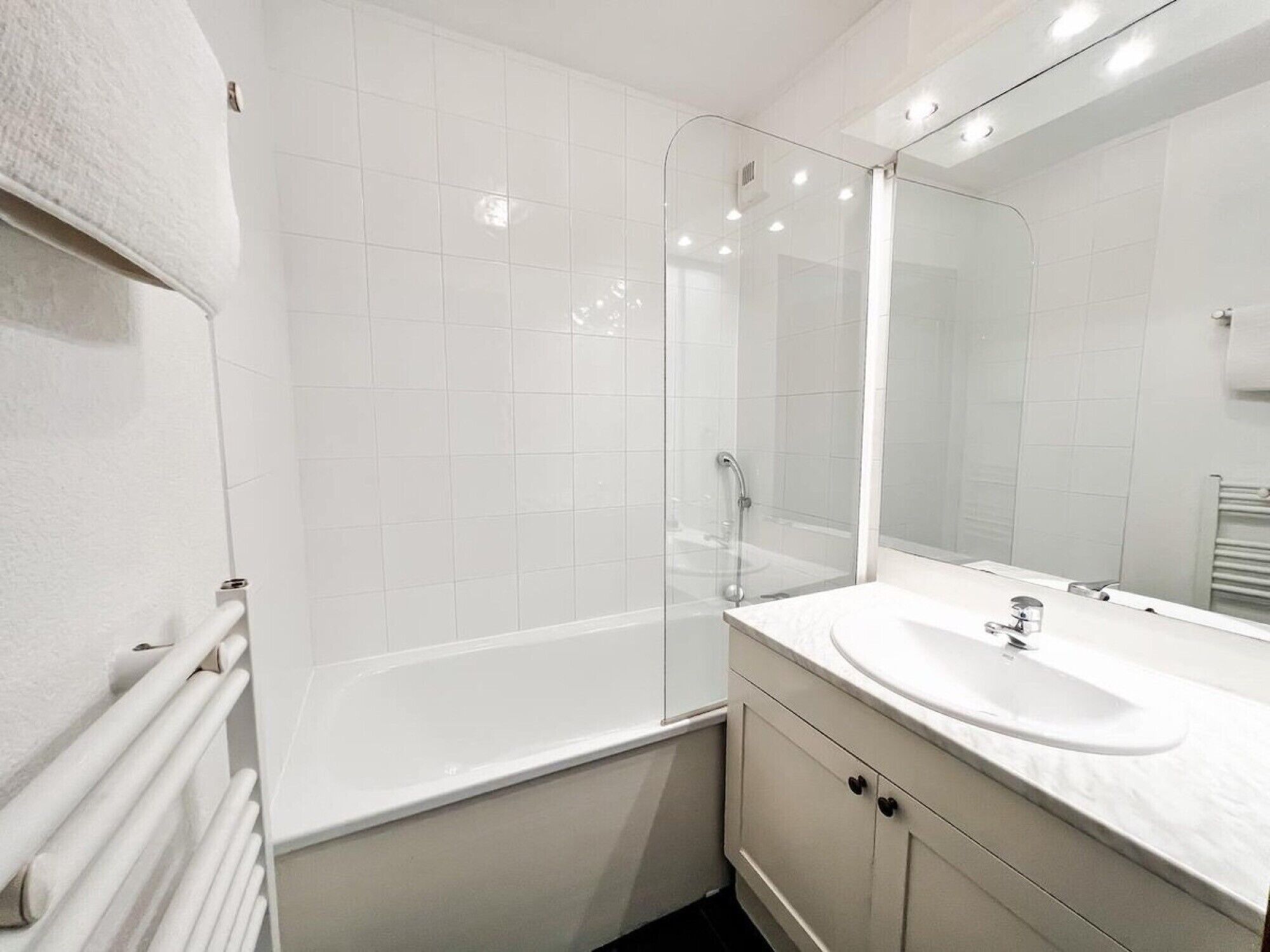 A modern bathroom with white tiles, a glass-enclosed shower, a white sink, and a large mirror.