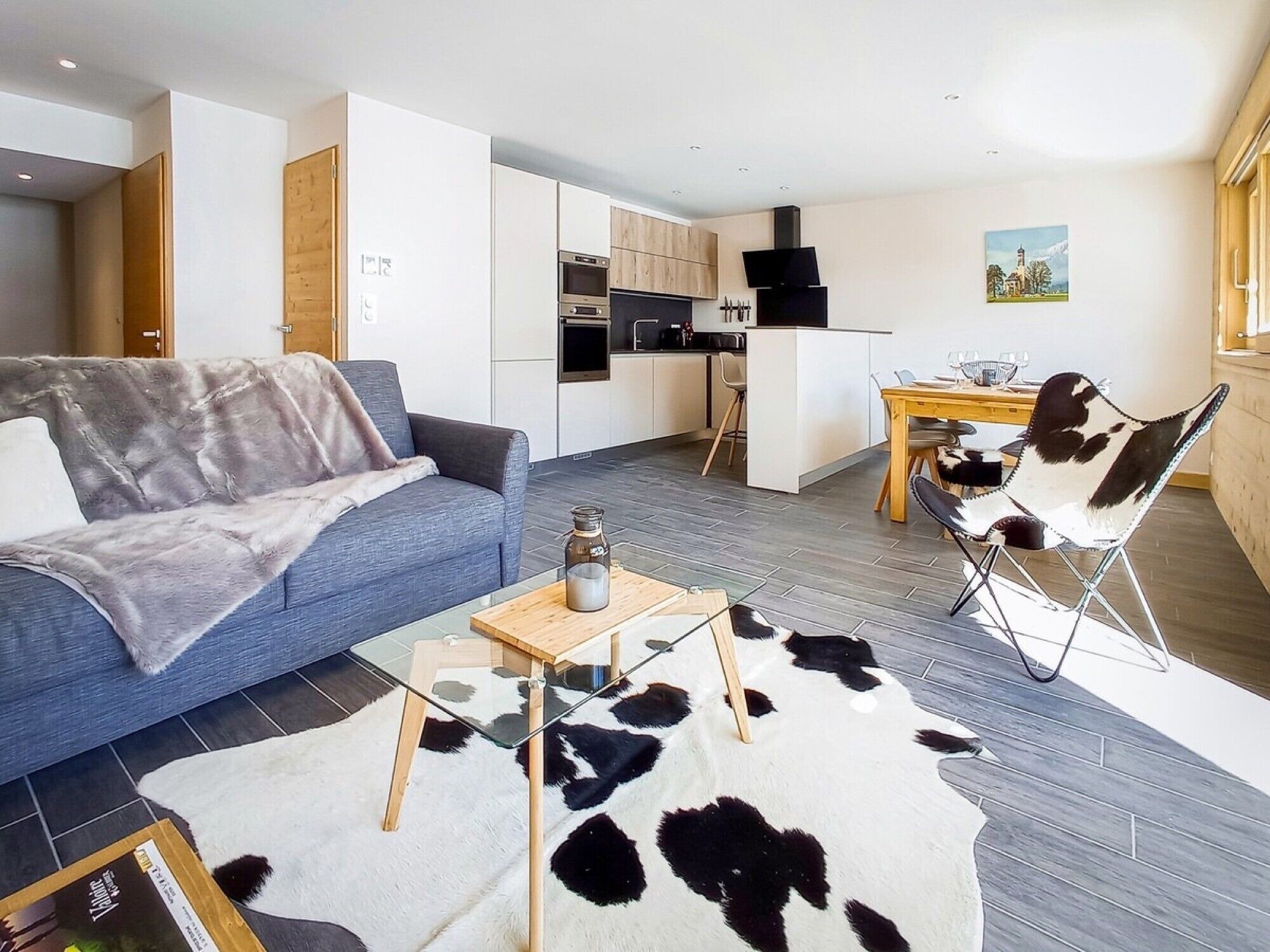 Una sala moderna con un sofá, una mesa de centro de vidrio, un tapete de cuero de vaca, una cocina con armarios blancos y un comedor con una silla.