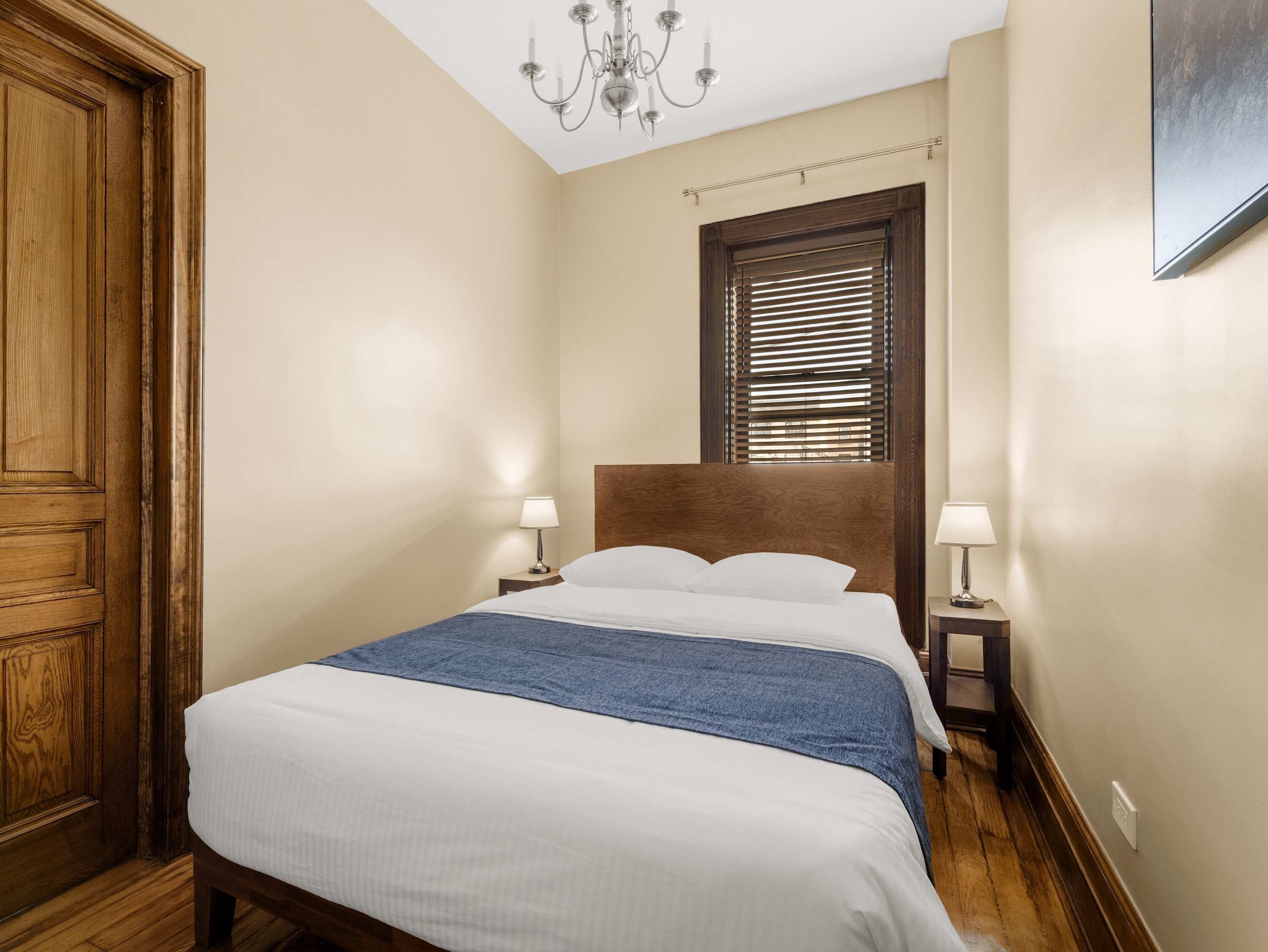 Une chambre d’hôtel avec un grand lit, une tête de lit en bois, deux tables de chevet avec des lampes, un lustre et une fenêtre avec des stores.