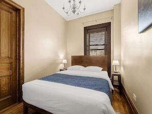 A hotel room with a large bed, wooden headboard, two bedside tables with lamps, a chandelier, and a window with blinds.