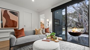 A modern living room with a grey sofa, a round ottoman, and a fireplace visible through a glass door. - Dinner Plain Mt Hotham Accommodation (Dinner Plain)