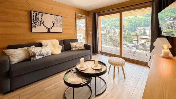 Een moderne woonkamer met een donkergrijze bank, een ronde salontafel en een klein krukje. Er is een groot raam met uitzicht op een balkon en groen buiten.