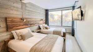 Une chambre avec deux lits, une tête de lit en bois, des lampes de chevet, une grande fenêtre et un téléviseur fixé au mur.