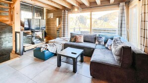 L’intérieur chaleureux d’un chalet avec un plafond en bois, un ensemble de canapés, un téléviseur et une grande fenêtre donnant sur le paysage extérieur.