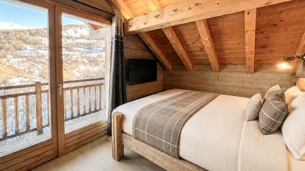 Une chambre avec un plafond en bois, un lit avec des oreillers, une télévision et une vue sur un paysage enneigé par une grande fenêtre.
