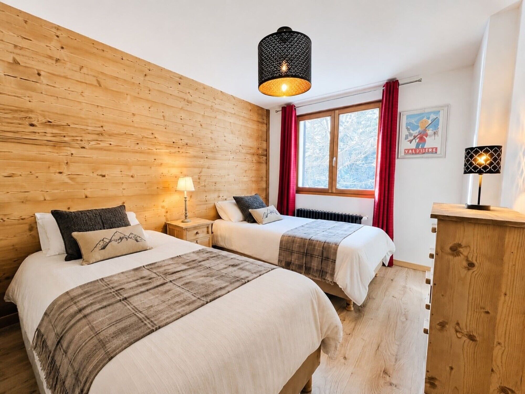 Una habitación con dos camas, paredes de madera, una ventana con cortinas rojas, un radiador y una lámpara colgante.