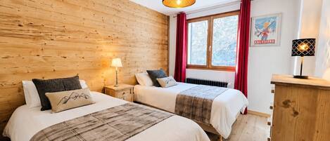 Una habitación con dos camas, paredes de madera, una ventana con cortinas rojas, un radiador y una lámpara colgante.