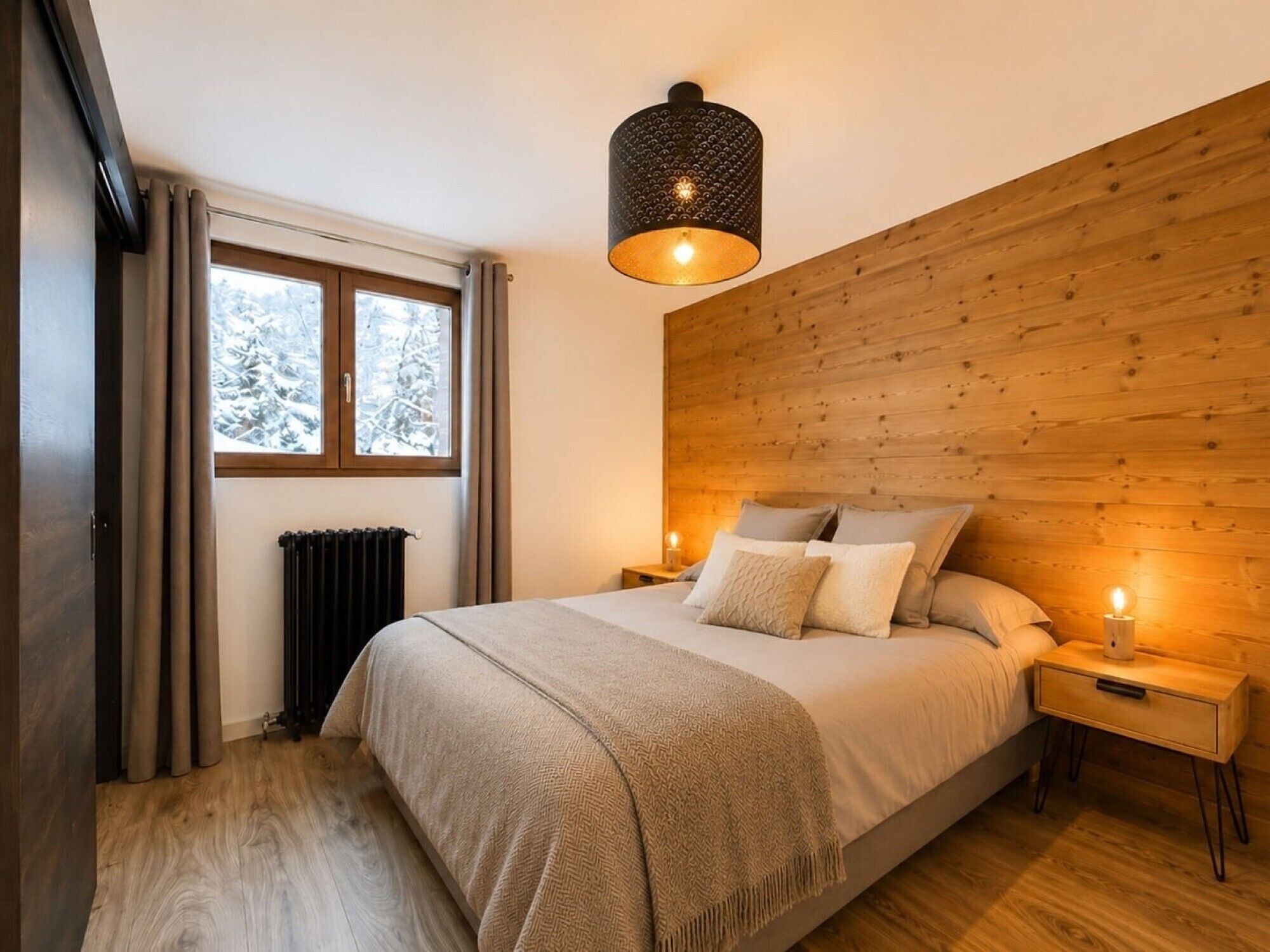 Une chambre à coucher avec une tête de lit en bois, un lit recouvert d’une couverture beige, deux lampes de chevet, une fenêtre avec des rideaux et un radiateur.