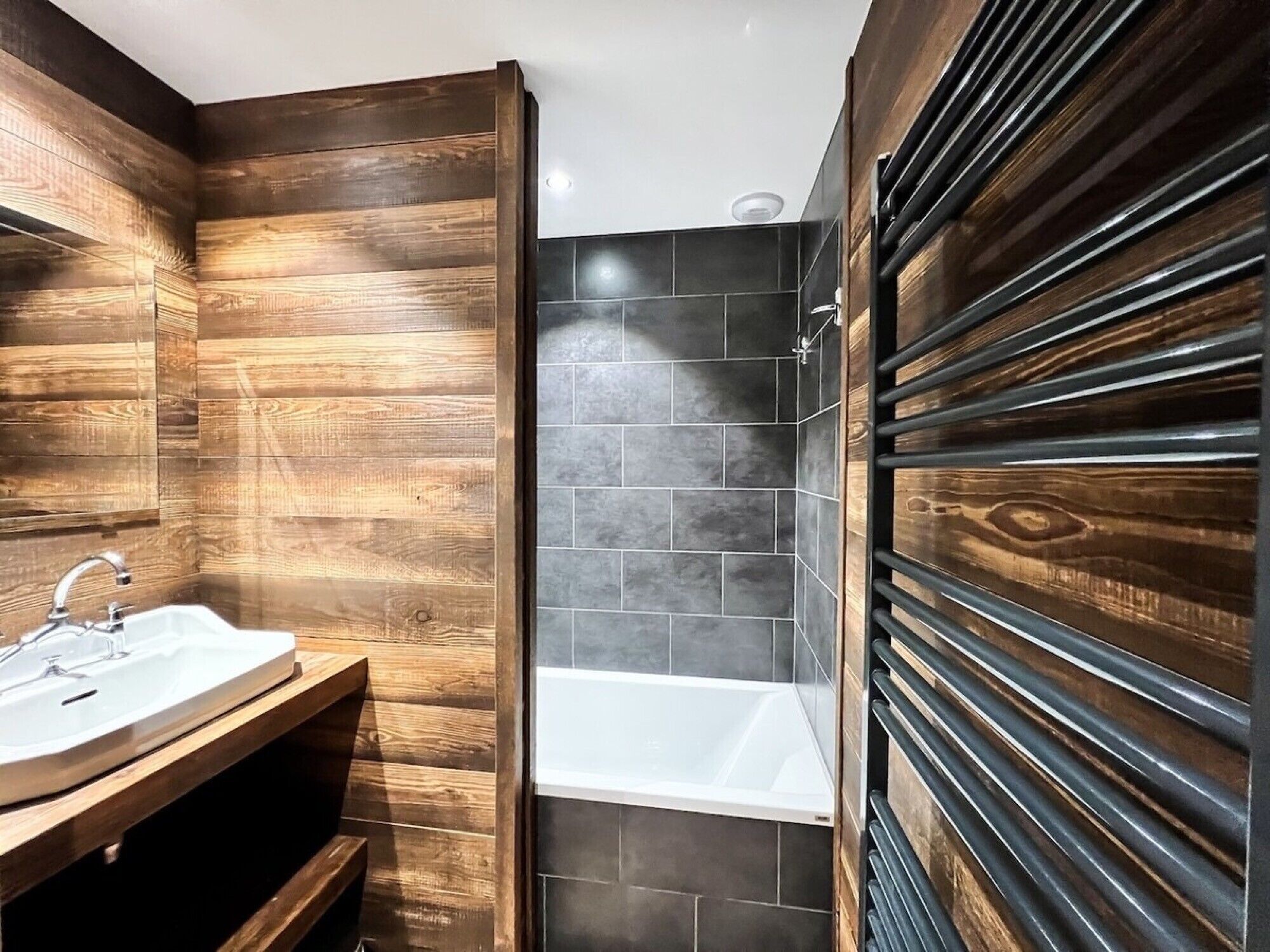 A bathroom with wooden paneling, a white sink, a bathtub, and dark tiled walls.