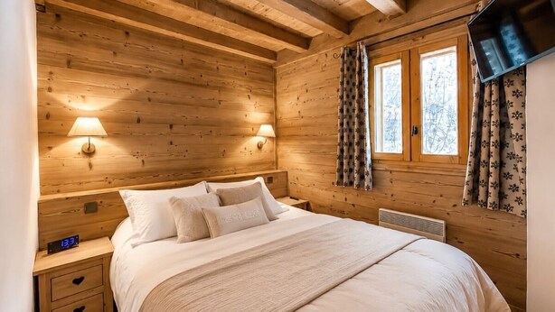 A wooden cabin bedroom with a bed, bedside lamps, a clock, and a TV mounted on the wall. - Apartment - Le Pti Chalet Valloire - 6 People (Valloire)
