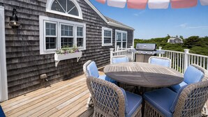 A deck with a round table and wicker chairs, a grill, and a white railing. - Longtail Cottage (Nantucket)