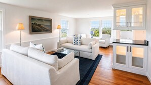 A modern living room with a sofa set, a coffee table, and a dining area with a kitchen in the background. - Longtail Cottage (Nantucket)