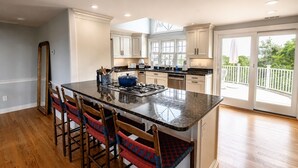 A spacious kitchen with a large island, wooden chairs, and a view of the outdoors through large windows. - Longtail Cottage (Nantucket)