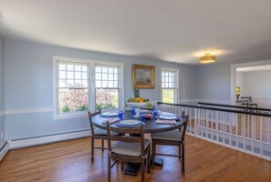 A dining area with a round table, chairs, and a fruit bowl. There are windows and a railing visible. - Longtail Cottage (Nantucket)