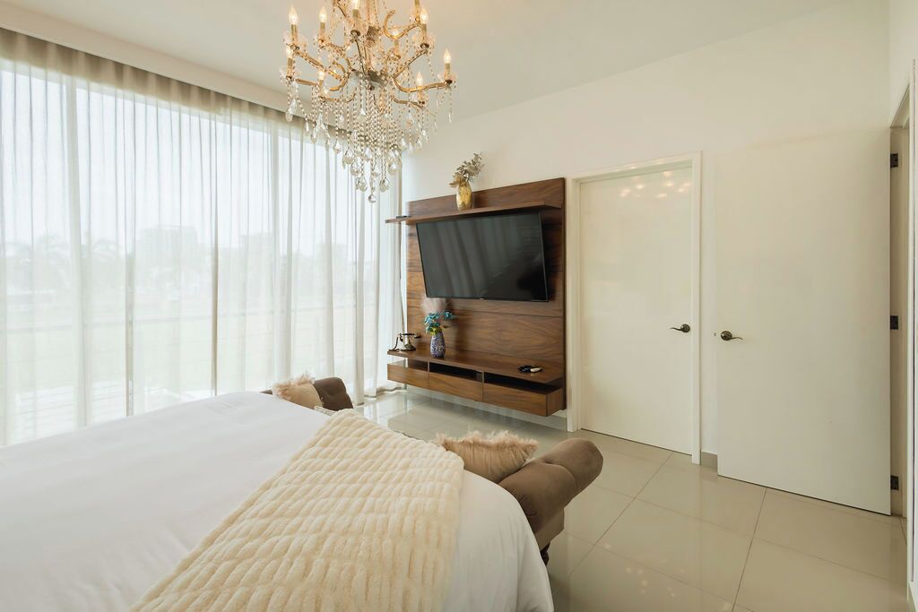 A modern bedroom with a large bed, a TV mounted on a wooden cabinet, a chandelier, and a view through large windows.