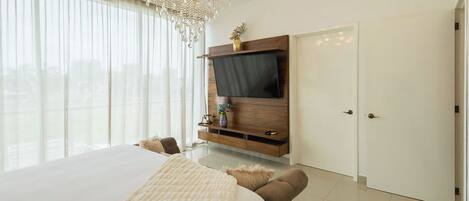 A modern bedroom with a large bed, a TV mounted on a wooden cabinet, a chandelier, and a view through large windows.