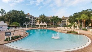 Uma piscina grande com uma fonte, rodeada por espreguiçadeiras e um edifício com vários andares e varandas.