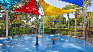 Ein Wasserspielbereich mit blauem Pool, umgeben von einem Zaun und beschattet durch farbenfrohe Sonnensegel.