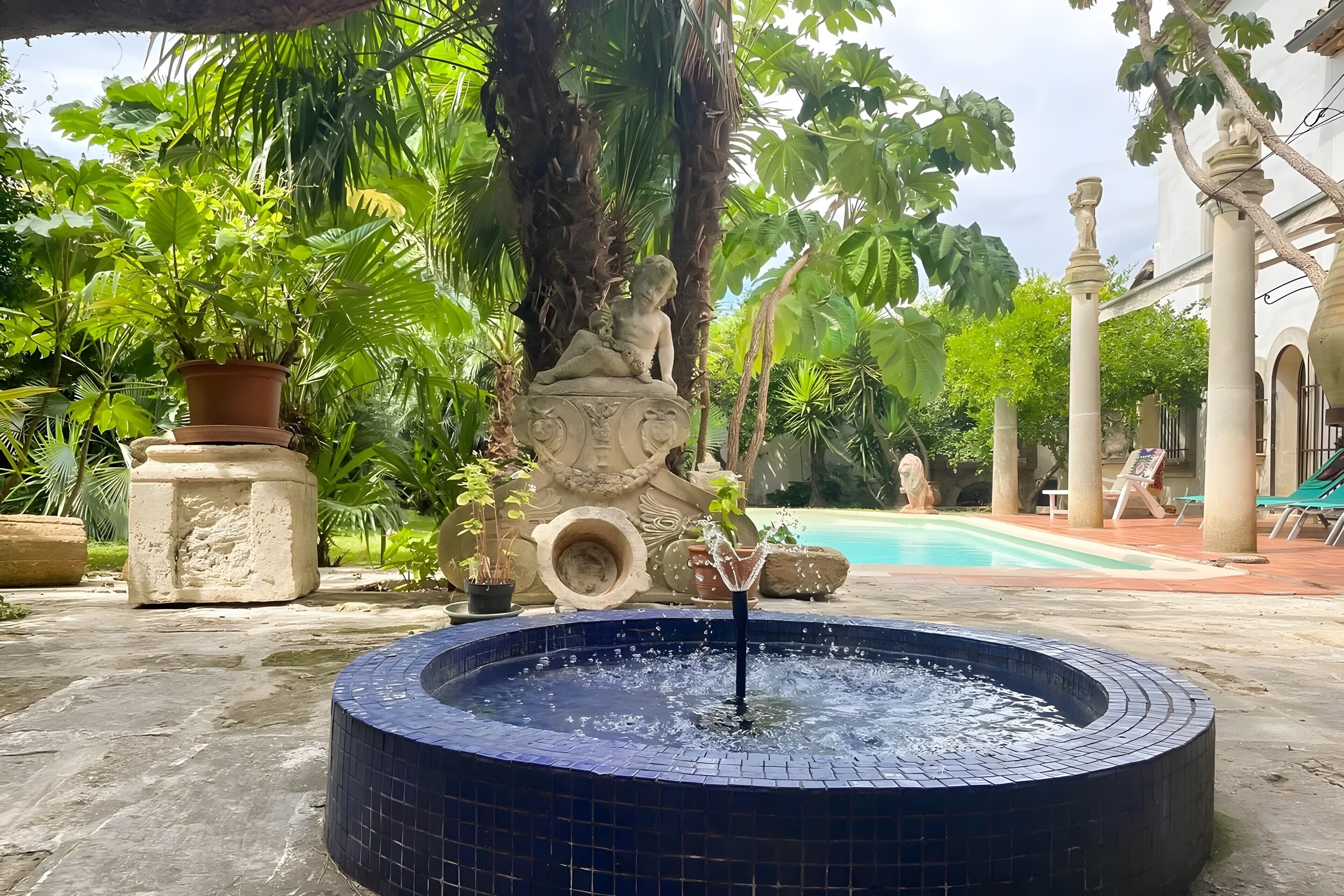 Ein Springbrunnen, aus dessen Mitte Wasser sprüht, umgeben von üppigem Grün und einem Pool im Hintergrund.