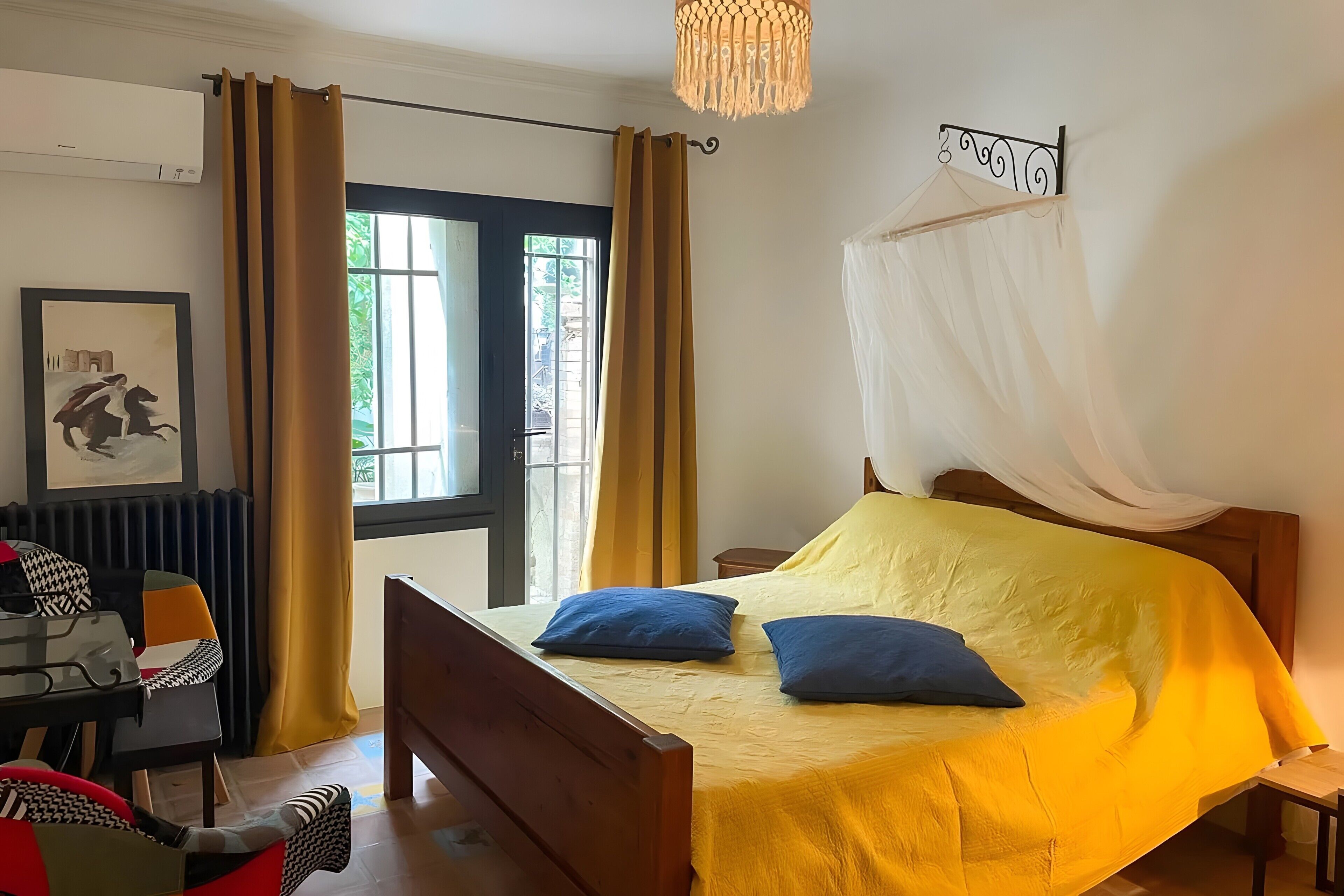 A room with a bed, a table with chairs, a chair, a picture frame, and a window with curtains.