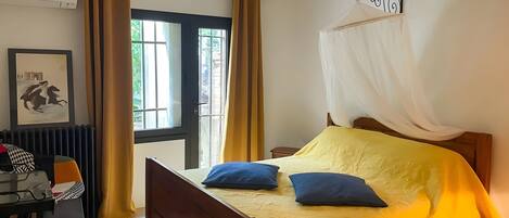A room with a bed, a table with chairs, a chair, a picture frame, and a window with curtains.