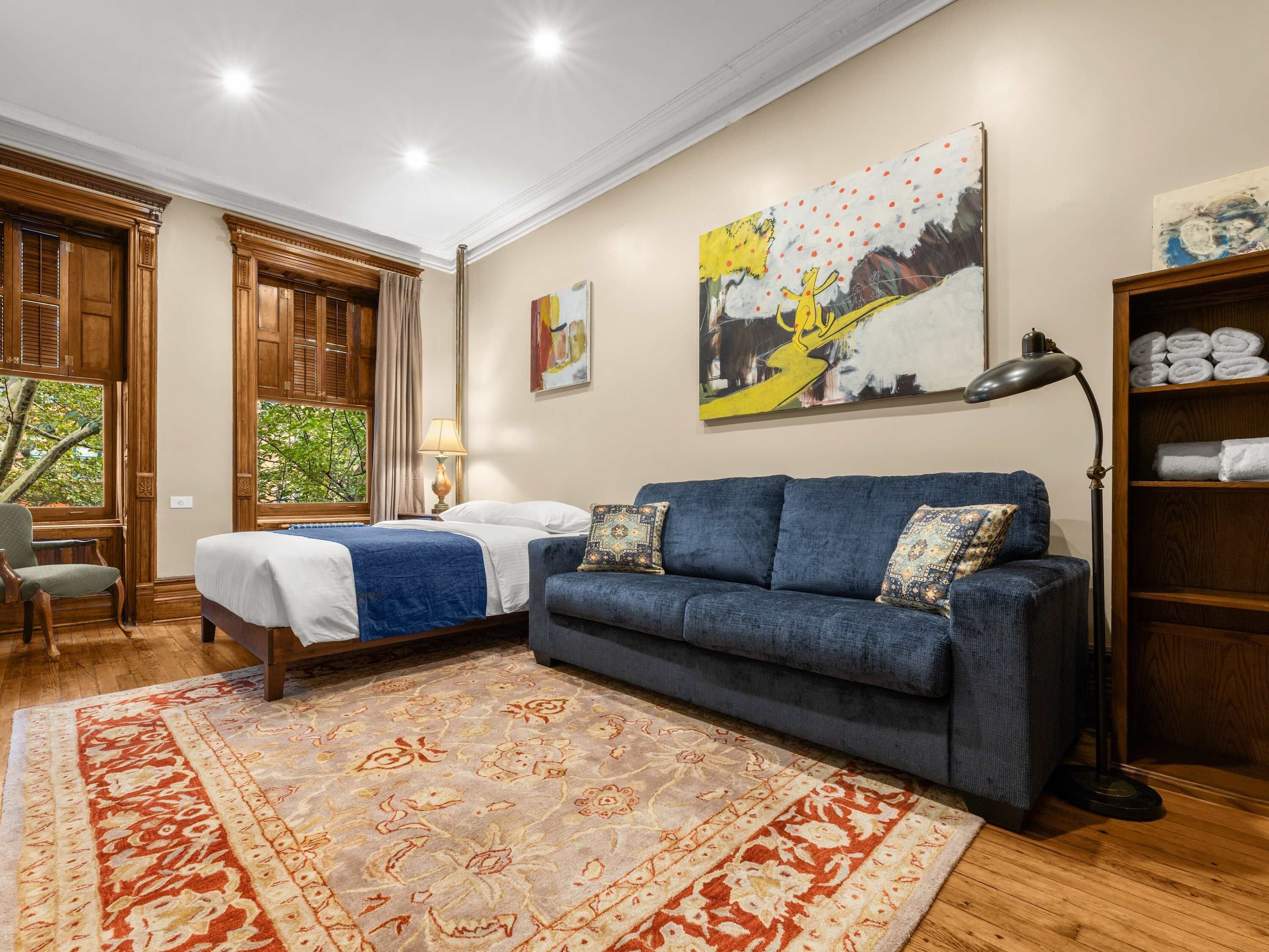 Habitación de hotel con cama, sofá azul, alfombra estampada, un cuadro y estante con toallas.