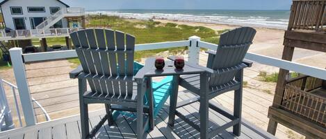 A deck with two Adirondack chairs and a table, overlooking a beach with a white building in the background.