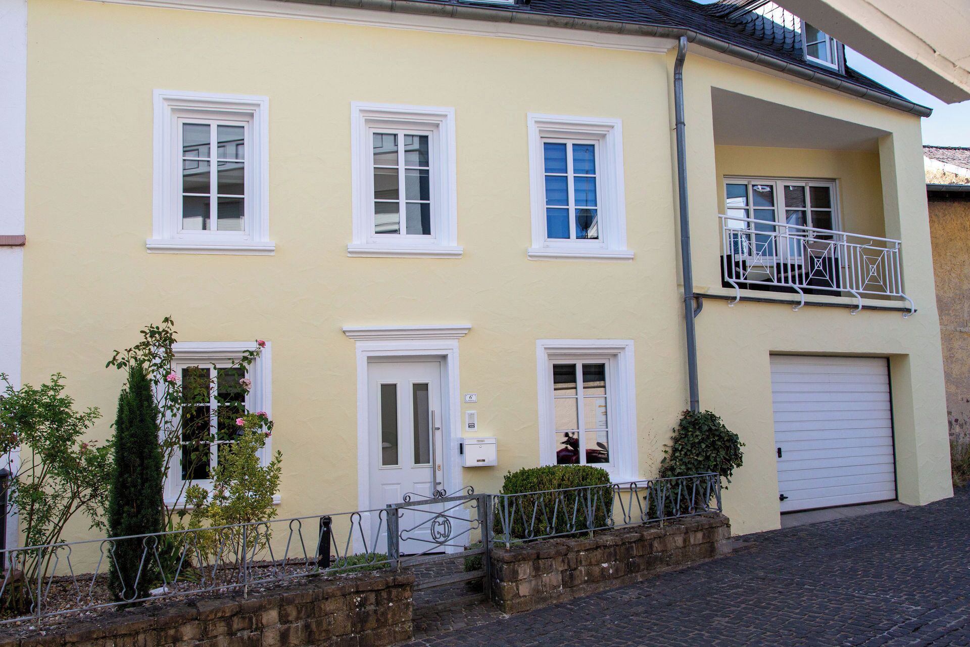 Ein zweistöckiges Gebäude mit gelber Fassade, weißen Fensterrahmen, einer sichtbaren Garagentür und einem kleinen Balkon mit Metallgeländer.