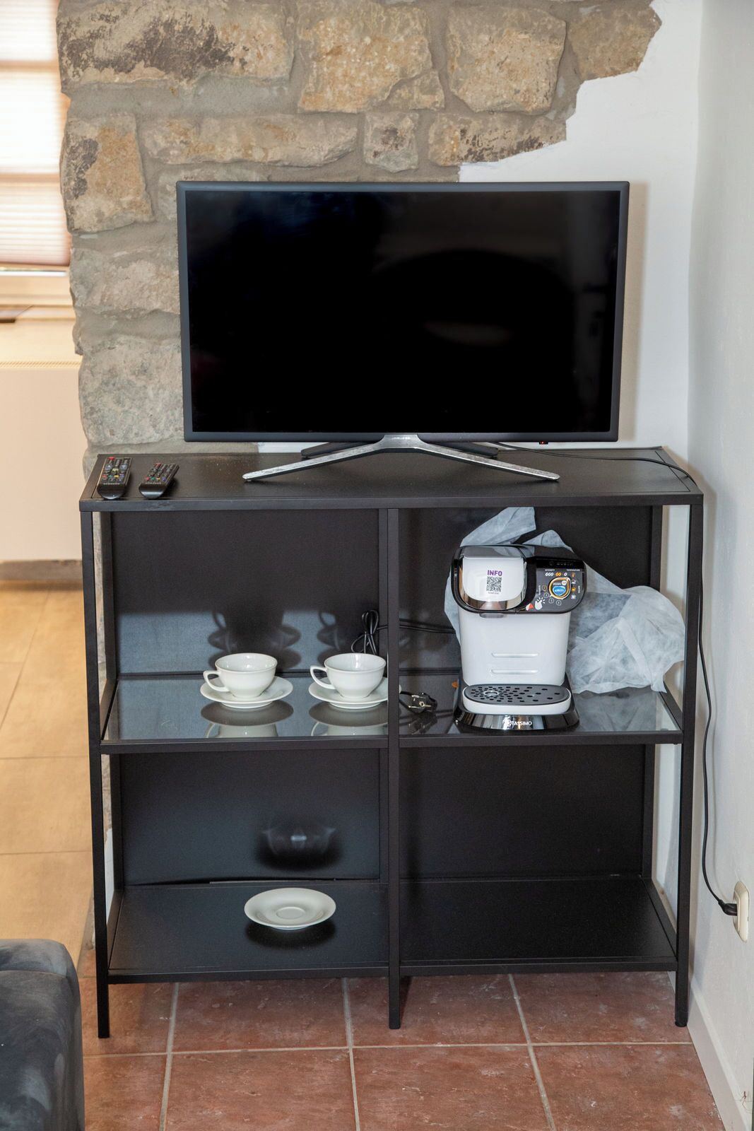 Un meuble de rangement noir pour appareils électroniques, avec un téléviseur à écran plat, des télécommandes, une tasse blanche, un bol, une assiette et une machine à café.