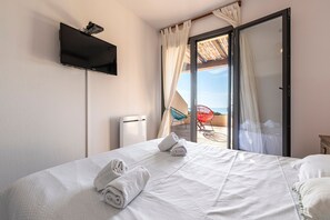 A hotel room with a bed, a flat-screen TV mounted on the wall, a sliding glass door leading to an outdoor area with a chair and umbrella. - Casa di Palombaggia - Appt avec belle vue mer (Porto-Vecchio)