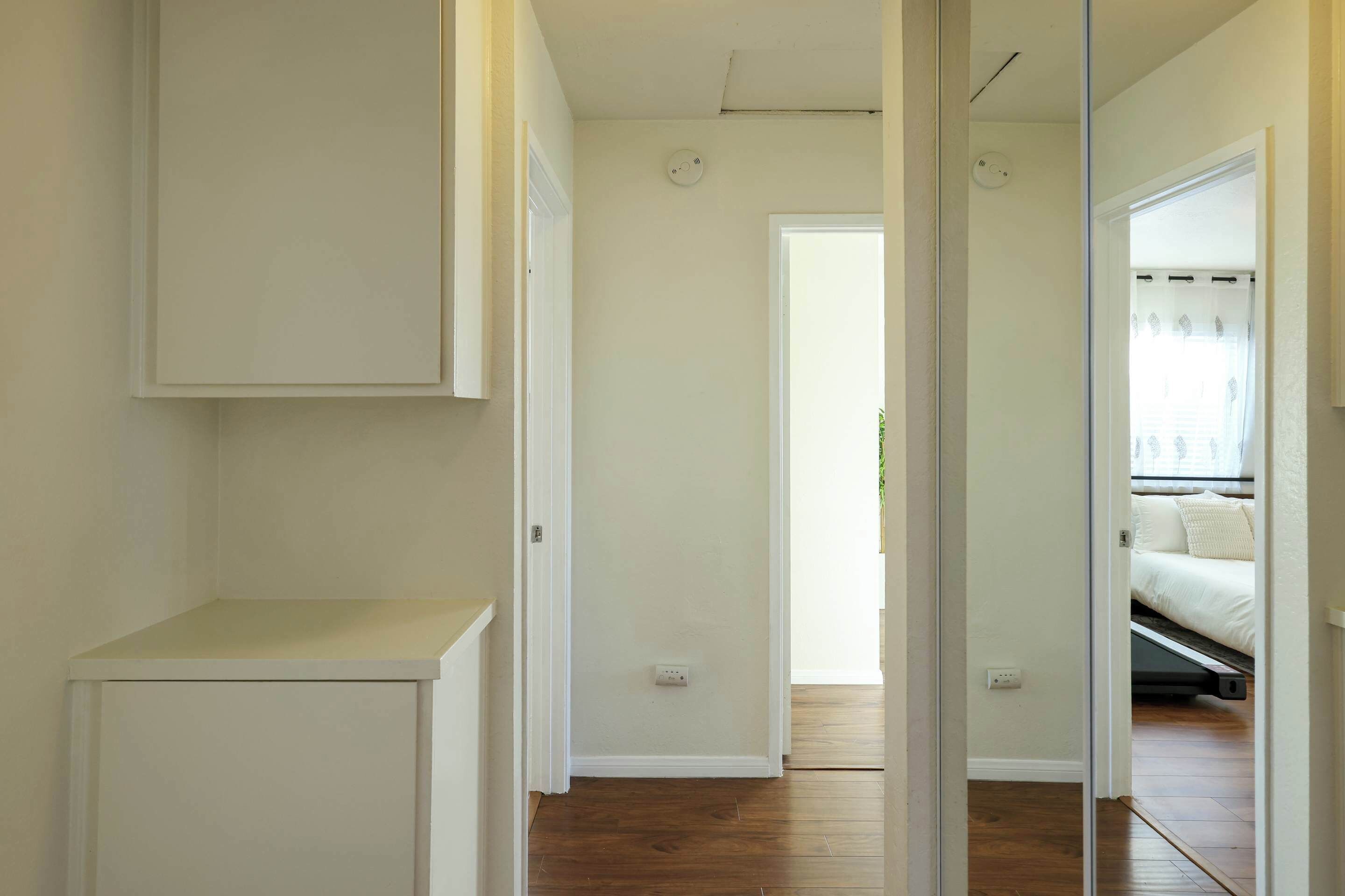 Een smalle gang met een witte kast, een deur naar een slaapkamer met een bed, en een zichtbare stopcontact op de muur.