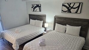Dos camas tamaño queen con ropa de cama blanca, cabeceros de madera y una mesita de noche con lámpara.