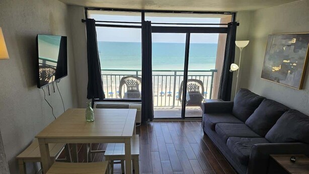 Una habitación con una mesa de madera, un sofá, un televisor y un balcón con vistas a la playa.