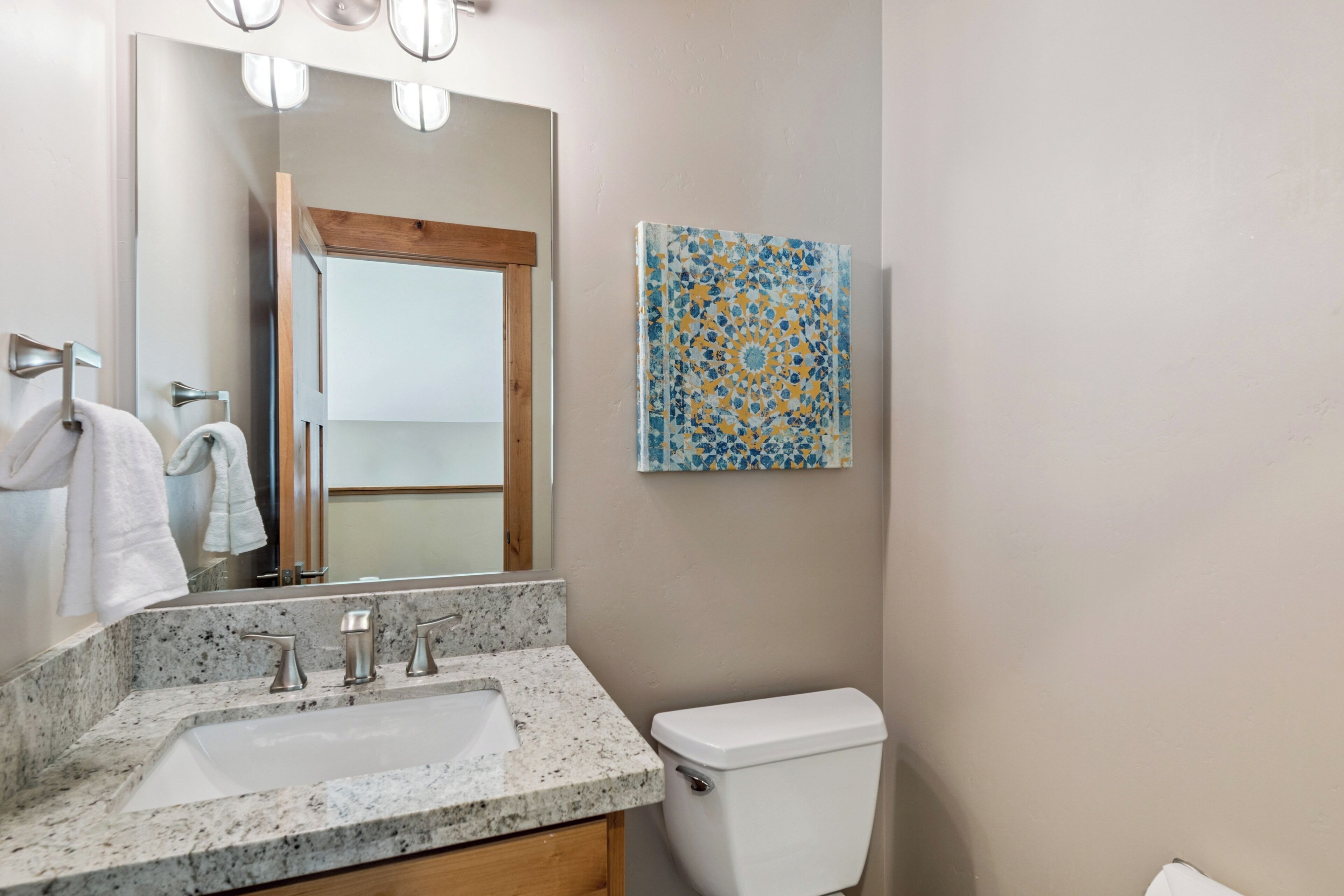 A bathroom with a granite countertop sink, a white toilet, a wooden door, and a decorative tile wall piece.