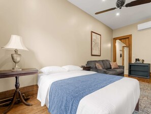 Ein Hotelzimmer mit einem Bett, einem Tisch mit einer Lampe, einer Couch, einem gerahmten Bild an der Wand und einem Deckenventilator.