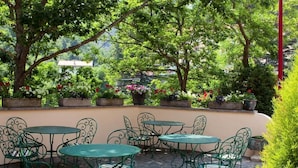 An outdoor patio with round tables and chairs, surrounded by lush greenery and flowering plants. - Hotel Val Du Tech  (Prats-de-Mollo-la-Preste)