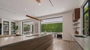 A modern kitchen with a large island, built-in cabinets, and a view of a green garden. - Villa Croft | Beverly Hills Luxury Hideaway (Los Angeles)