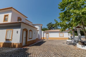 Un patio con pavimento de adoquines, edificios blancos con detalles en naranja y un árbol que da sombra.
