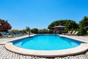 Una piscina con sillas de descanso y sombrillas, rodeada de árboles y un cielo despejado.