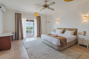 Un dormitorio con cama, mesitas de noche, ventilador de techo, ventana con cortinas y vista a zonas verdes.