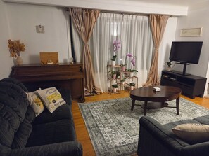 Un salon comprenant un canapé foncé, une table basse, un meuble pour la télévision, un piano et un support pour plantes.