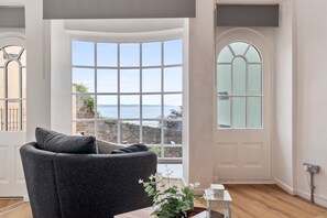 A room with a curved bay window, a grey sofa, a wooden floor, and a view of the sea. - Tenby Harbour View - Heart of Tenby, Sea Views (Tenby)