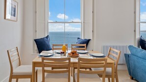 Un coin repas avec une table dressée pour quatre personnes, une vue sur la mer par une grande fenêtre et un canapé bleu dans un coin.