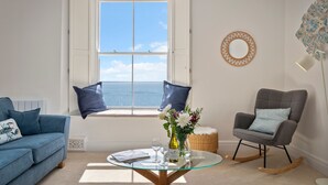 Une pièce avec vue sur la mer, un canapé bleu, une table en verre ornée d’un vase de fleurs et un fauteuil à bascule gris.