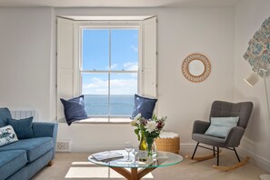 Ein Zimmer mit Meerblick, ein blaues Sofa, ein Glastisch mit Blumenvase und ein grauer Schaukelstuhl.