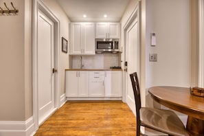 A compact kitchen with white cabinetry, a stainless steel microwave, and a wooden dining table.